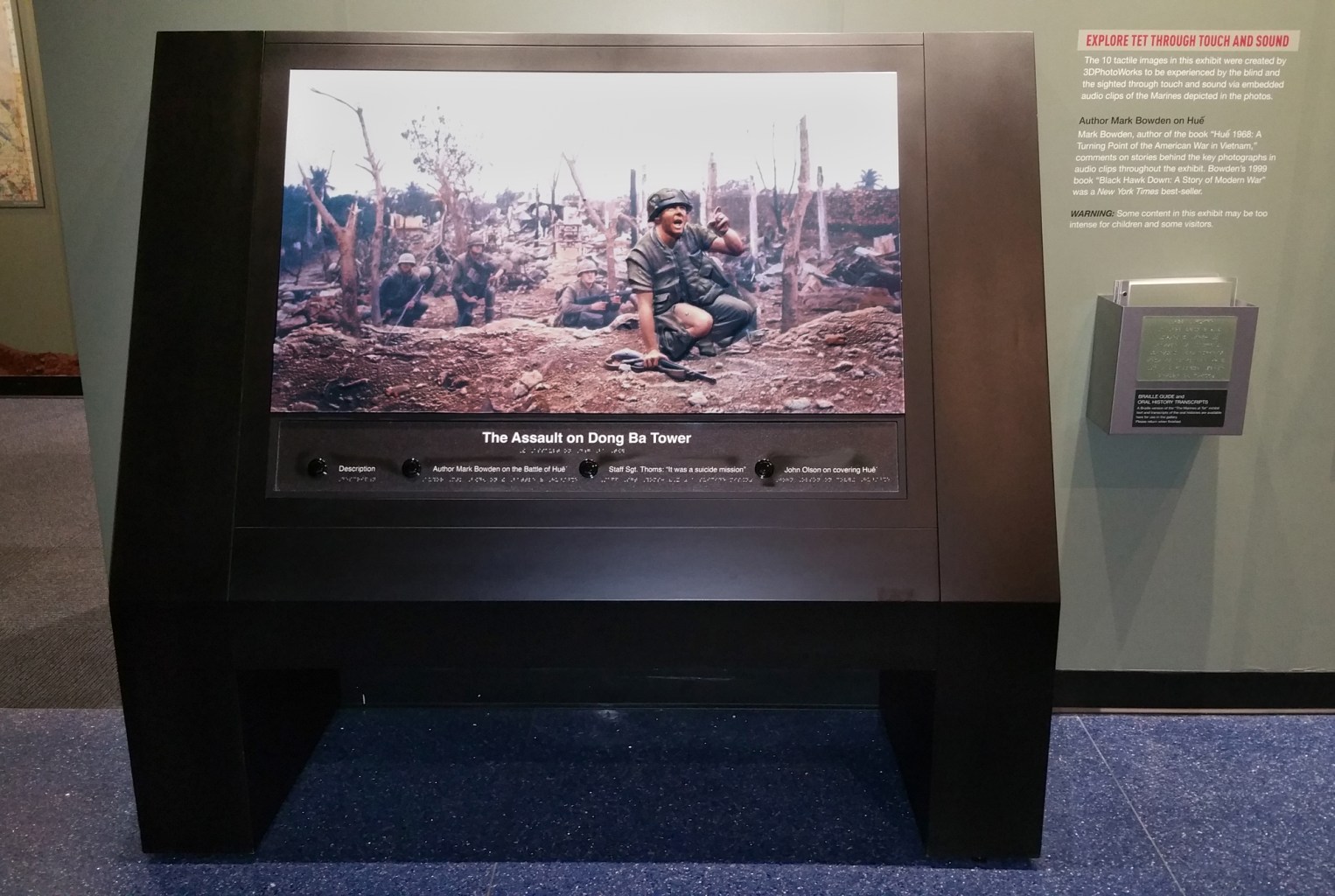 A tactile print shows armed soldiers crouching in a field. The text, reproduced in braille, reads "The Assault on Dong Ba Tower," with buttons that say "Author Mark Bowden on the Battle of Hue," "Staff Sgt. Thoms: 'It was a suicide mission,'" and "John Olson on covering Hue."