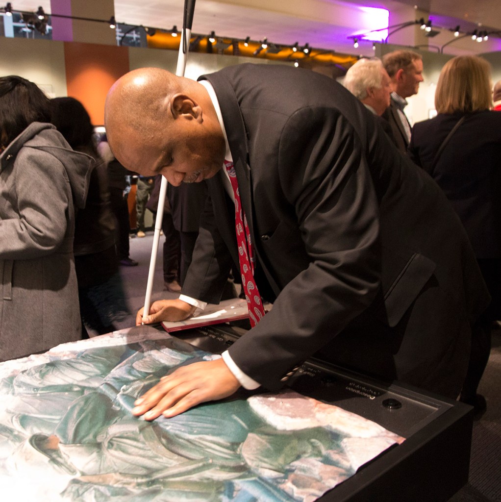 A man holding a cane leans over a tactile print on a table with his hand placed on its raised surface.
