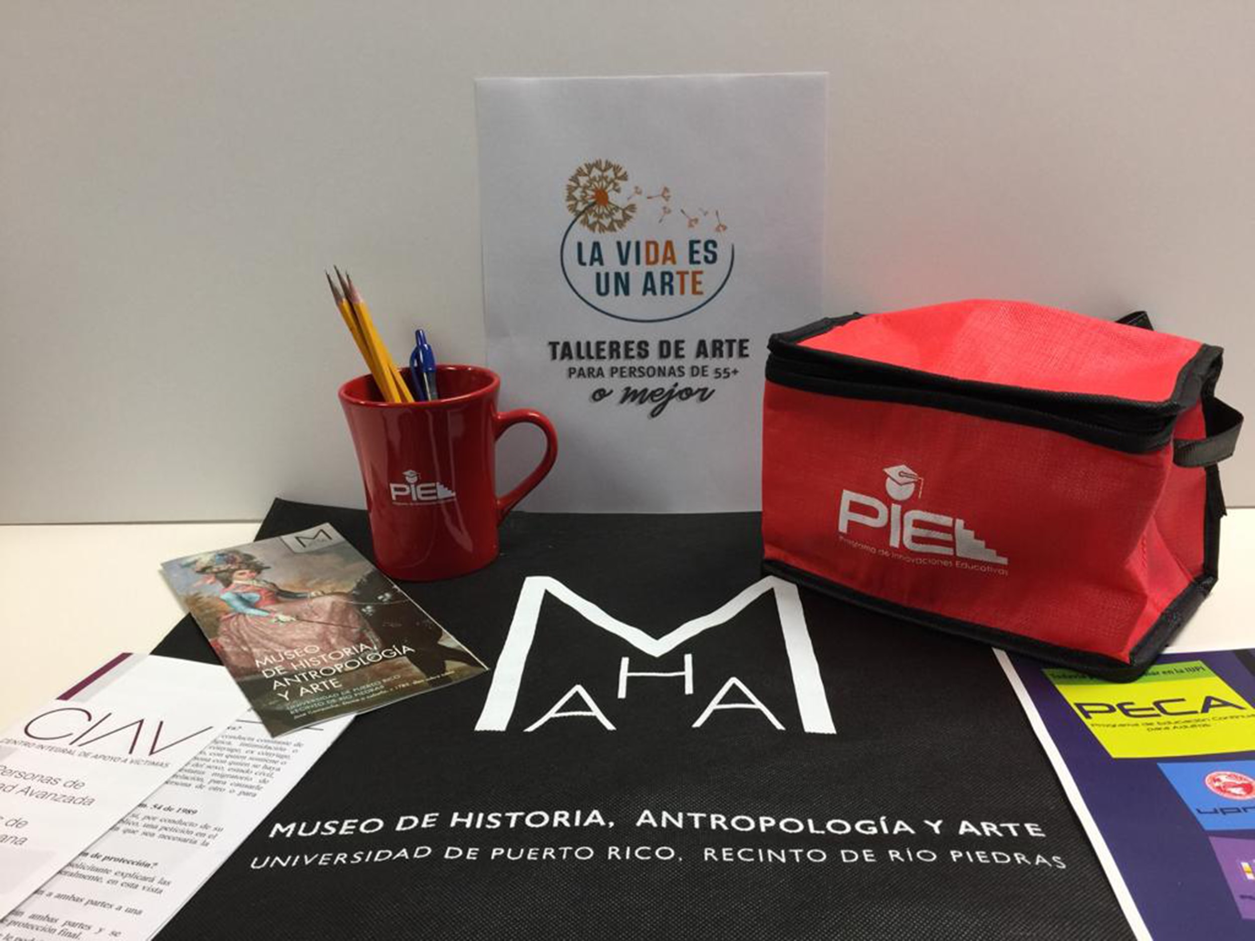 A sign for the program reads "La vida es un arte / Talleres de arte para personas de 55+ o mejor." Underneath is a a mug and lunchbox filled with goodies distributed to participants.