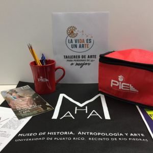 A sign for the program reads "La vida es un arte / Talleres de arte para personas de 55+ o mejor." Underneath is a a mug and lunchbox filled with goodies distributed to participants.