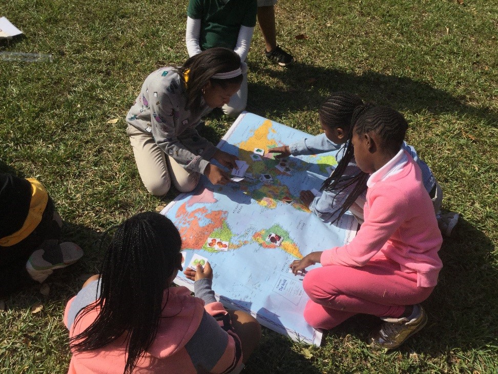 A group of children sits around a map of the world in the grass, with loose pieces of paper with foods on them. They place them on sections of the map.