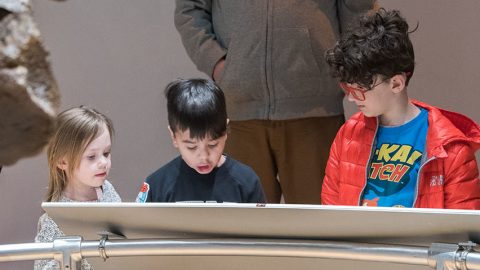 Three children gather around a display table inside a museum, while an adult stands behind them.