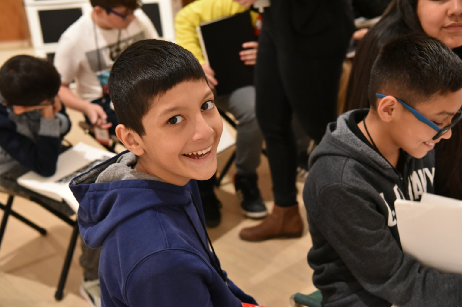 One student smiles at the camera, as others sit around working on an activity.