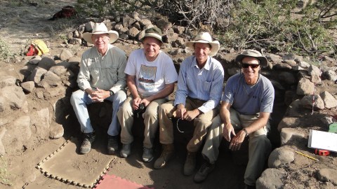 Four team members pose sitting in a recess in the earth surrounded by stone foundation.