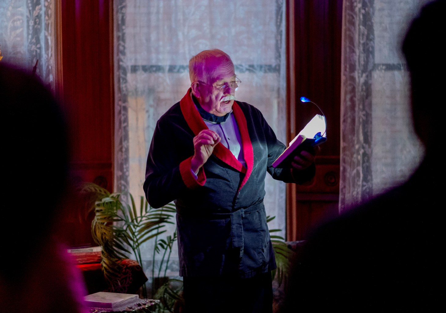 An actor stands in a dimly lit interior of the house museum holding a book, while audience members watch.