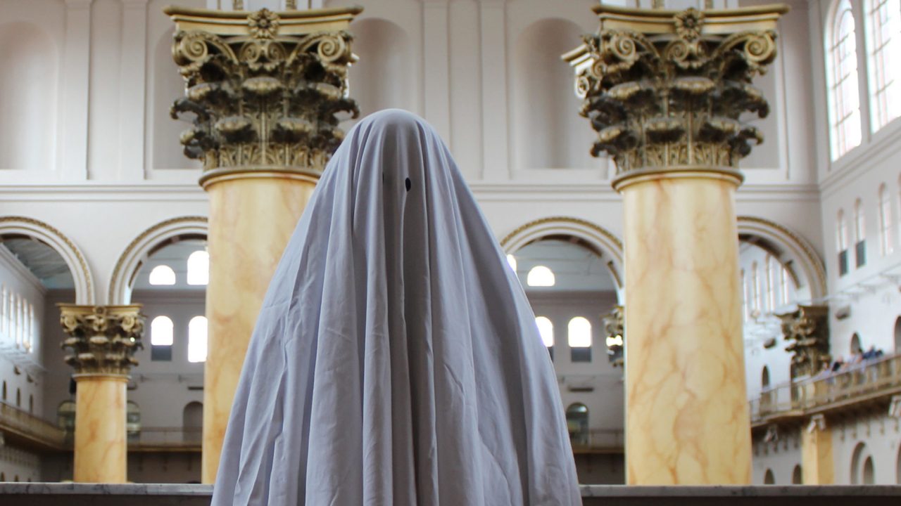 A sheet-with-eye-holes ghost stands in the middle of a historic museum building.