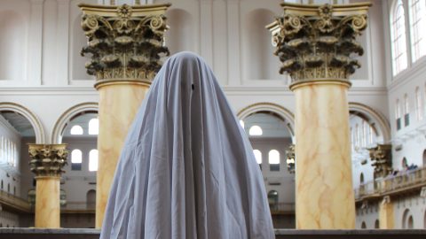 A sheet-with-eye-holes ghost stands in the middle of a historic museum building.