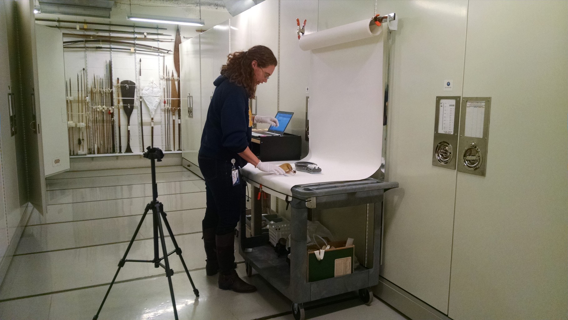 A museum employee stands at an imaging station with an object laid out on a white sheet of paper, and a camera pointed at it.