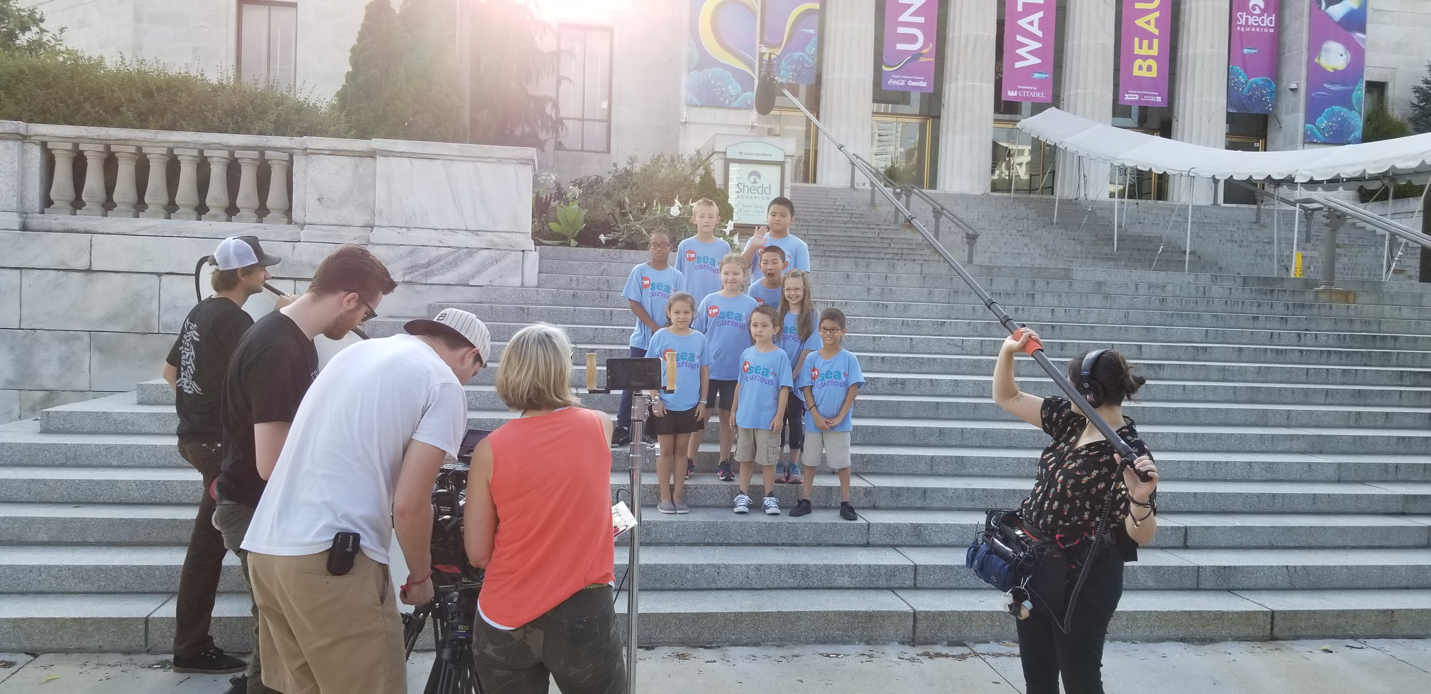 A film crew records a group of children on the aquarium's steps.