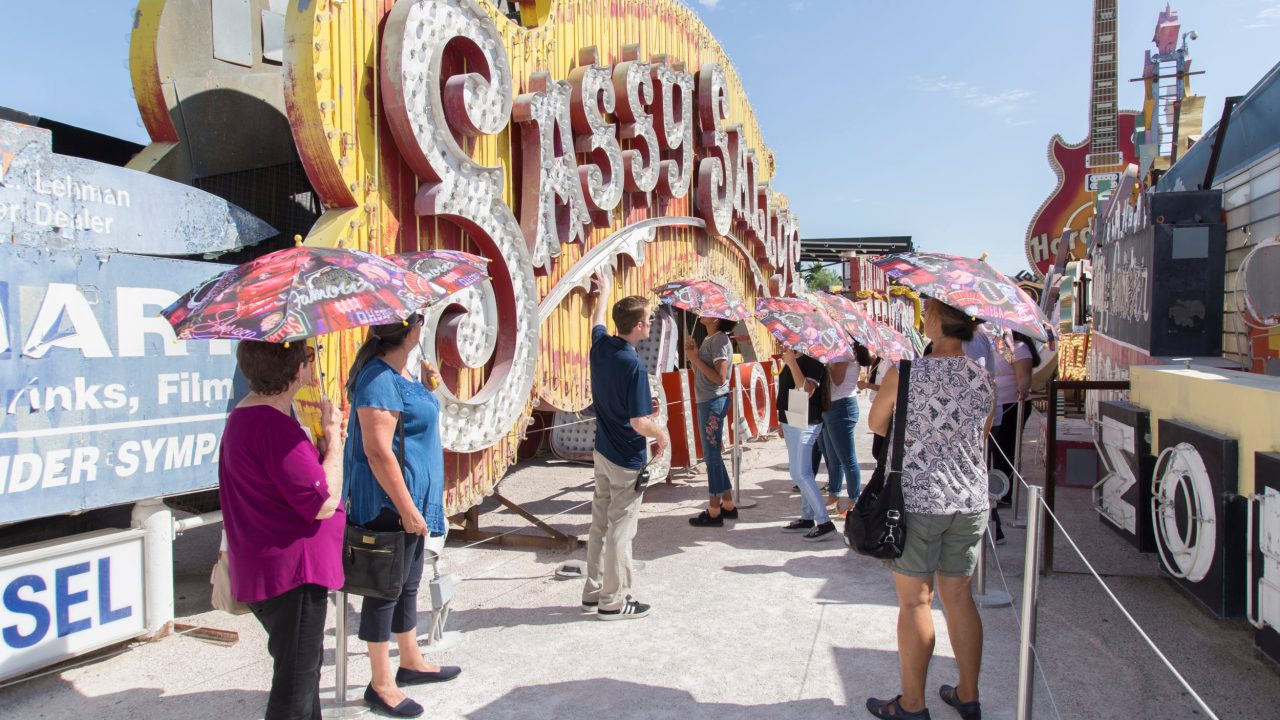 A group of people holding parasols stands outdoors in front of a large neon sign that says "Sassy."