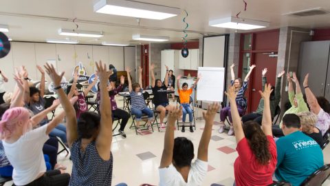 A group of people sitting in a circle all lifting their hands over their heads at the same time.