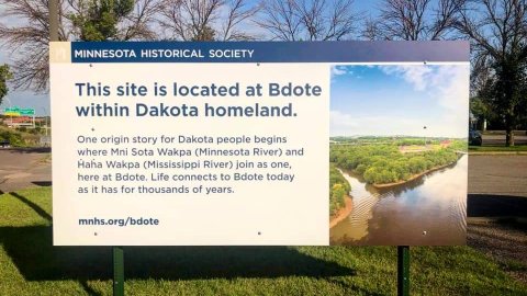An outdoor sign that reads "This site is located at Bdote within Dakota homeland. One origin story for Dakota people begins where Mni Sota Wakpa (Minnesota River) and Haha Wakpa (Mississippi River) join as one, here at Bdote. Life connects to Bdote today as it has for thousands of years.