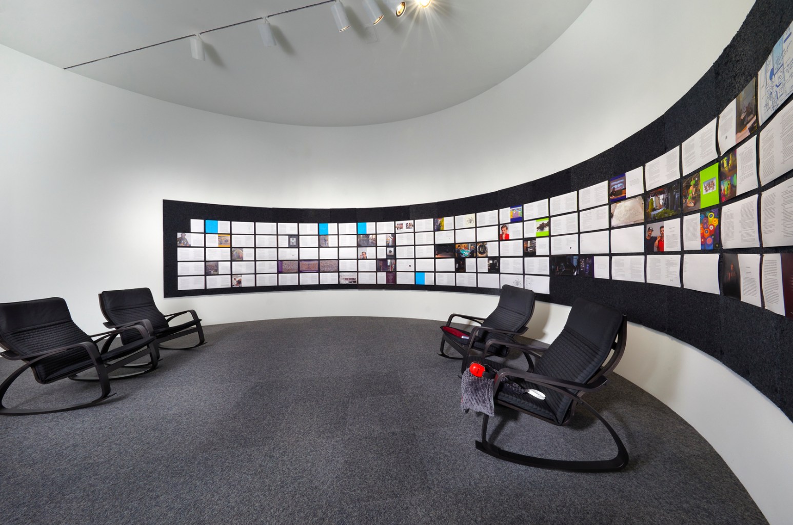 In a room with curved walls, rocking chairs are arranged in front of a grid of images.