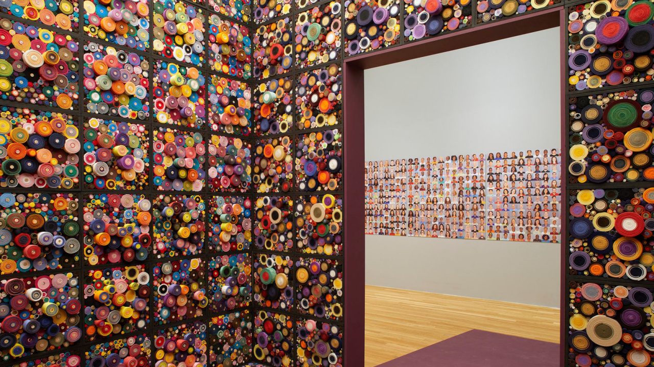 An immersion room inside of a museum gallery has cubes of textured, colorful bundles around the walls, and a grid of portraits is visible in the gallery beyond the door.