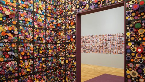 An immersion room inside of a museum gallery has cubes of textured, colorful bundles around the walls, and a grid of portraits is visible in the gallery beyond the door.