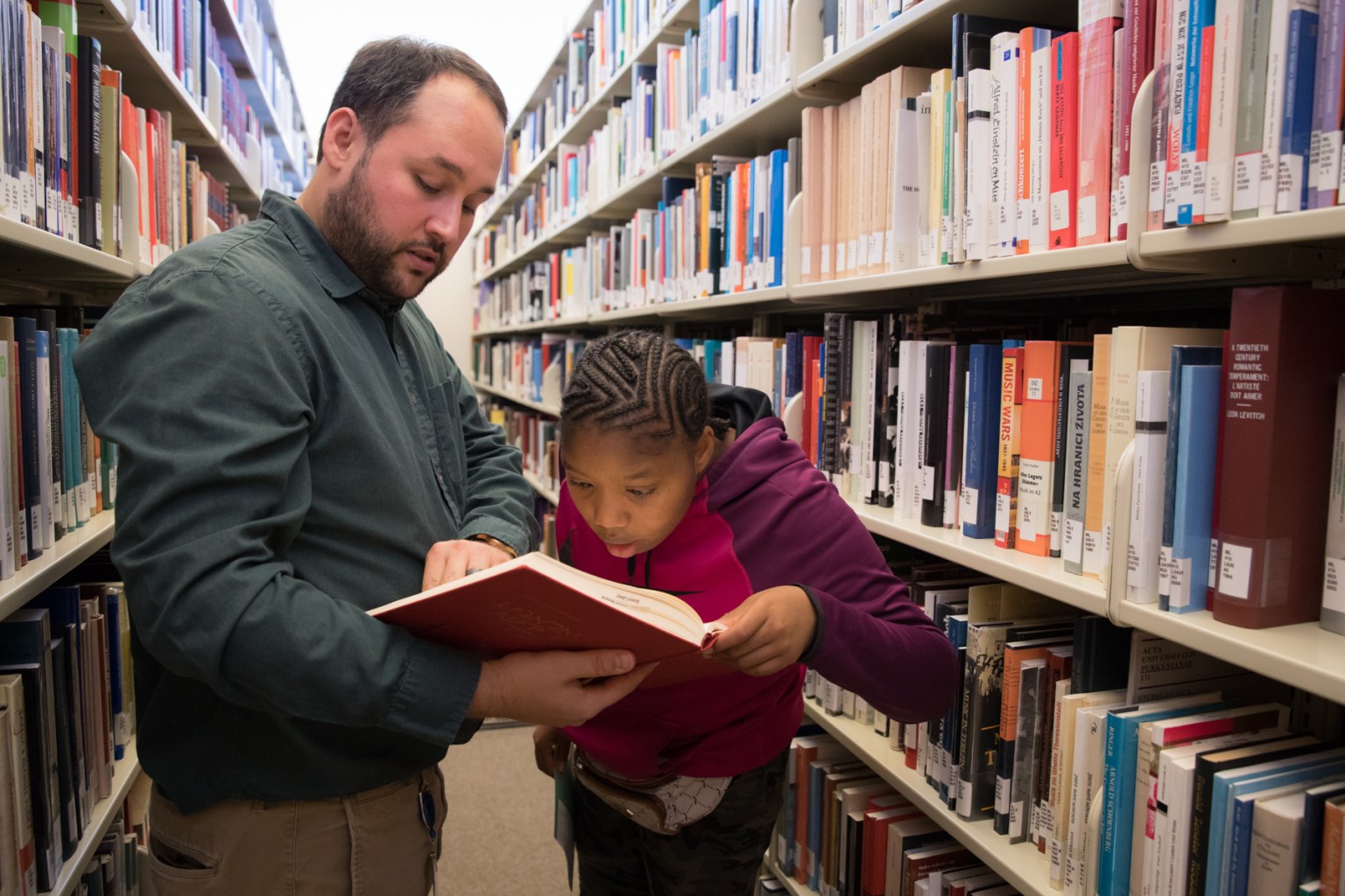 A staff person stands in library stacks with a student, holding a book open for her to read.