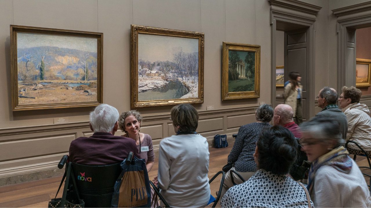 In a museum gallery in front of paintings, an educator kneels down talking to a group of program participants, one of whom is seated in a wheelchair.