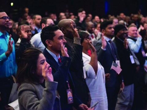 A large group of people stand with their hands raised in a naturalization ceremony.
