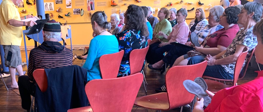 Seated participants in a yellow room listen to a standing speaker.