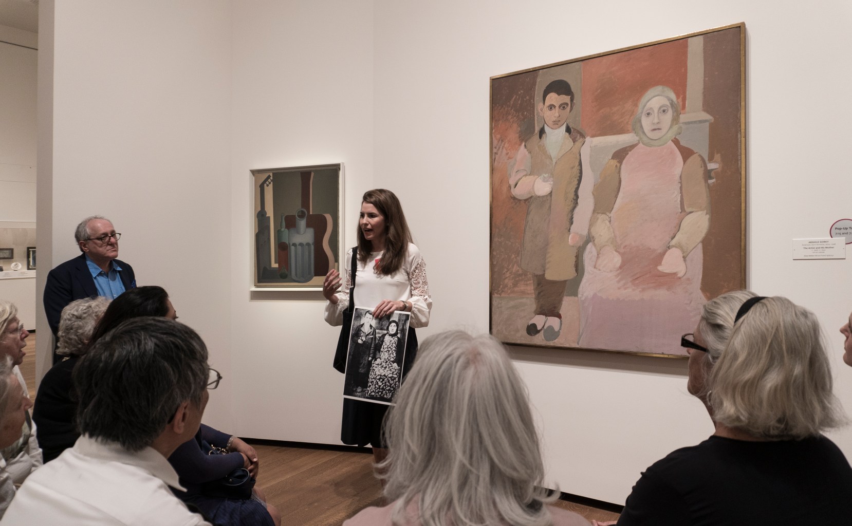 An educator stands in front of a painting addressing a seated group of participants. The painting behind her shows a standing child and a seated adult, and is rendered in flat blocks of earth-toned color. The educator holds up a print-out of a photograph of the subjects in real life.