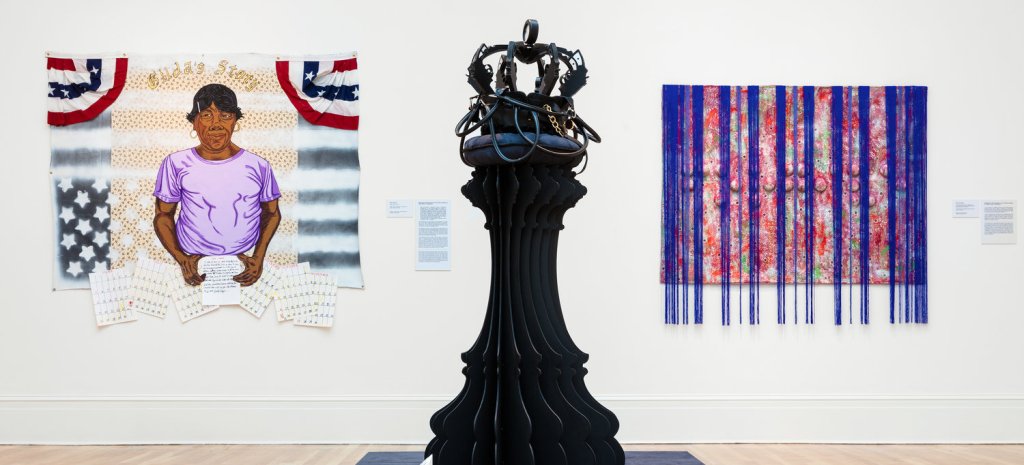 Three pieces of art in a museum gallery: one portrait of a subject in front of an American flag, one canvas with blue fringe hanging from it, and one large sculpture resembling a chess piece