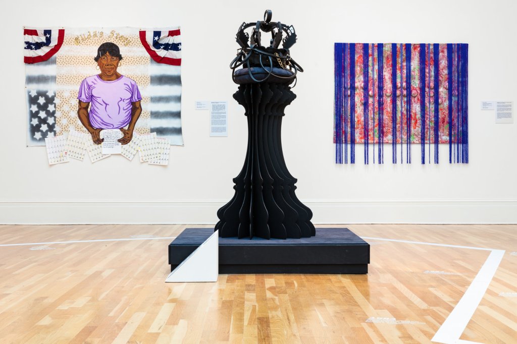 Three pieces of art in a museum gallery: one portrait of a subject in front of an American flag, one canvas with blue fringe hanging from it, and one large sculpture resembling a chess piece