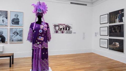 Artworks in a museum gallery, including photographed portraits and a purple dress and feathered headpiece on a mannequin