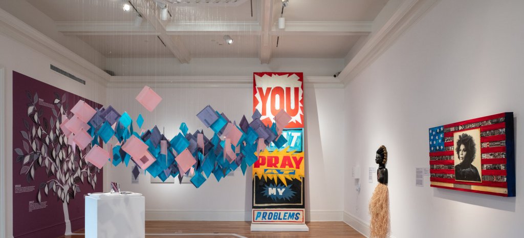 A variety of artworks in a museum gallery: pieces of paper suspended from the ceiling, a large drawing of a tree, and a piece that says "You can't pray at my problems" in bold lettering. A timeline in white font is visible on the wood floor.