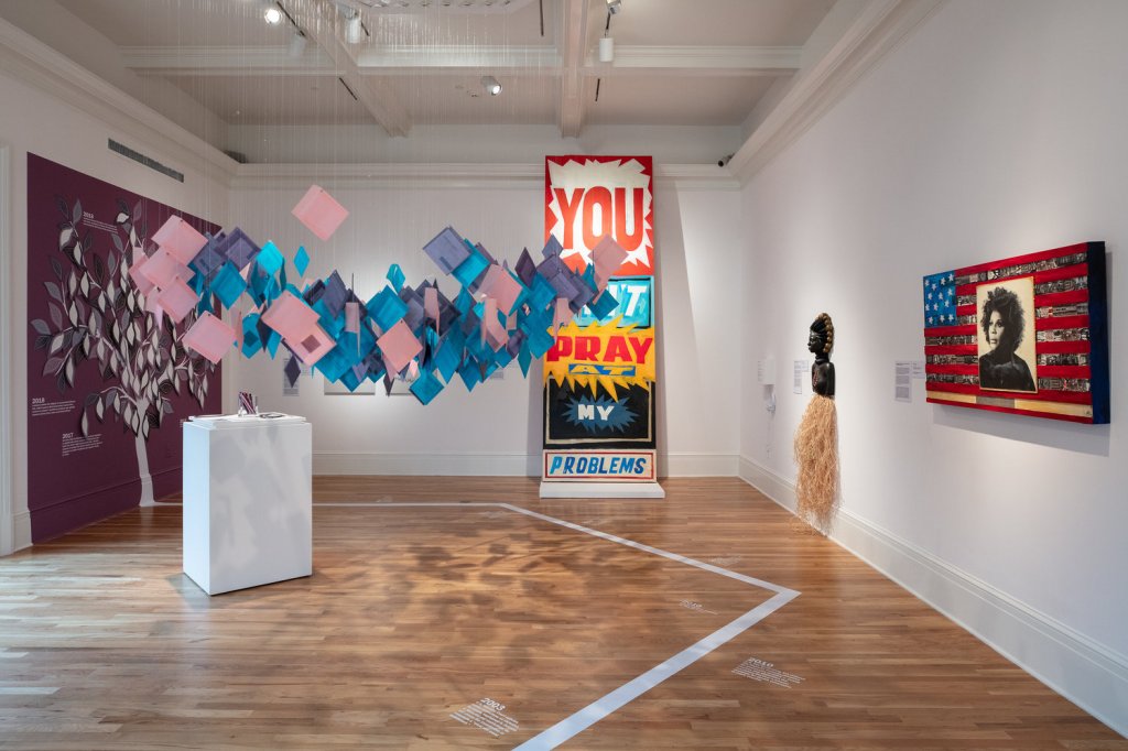 A variety of artworks in a museum gallery: pieces of paper suspended from the ceiling, a large drawing of a tree, and a piece that says "You can't pray at my problems" in bold lettering. A timeline in white font is visible on the wood floor.