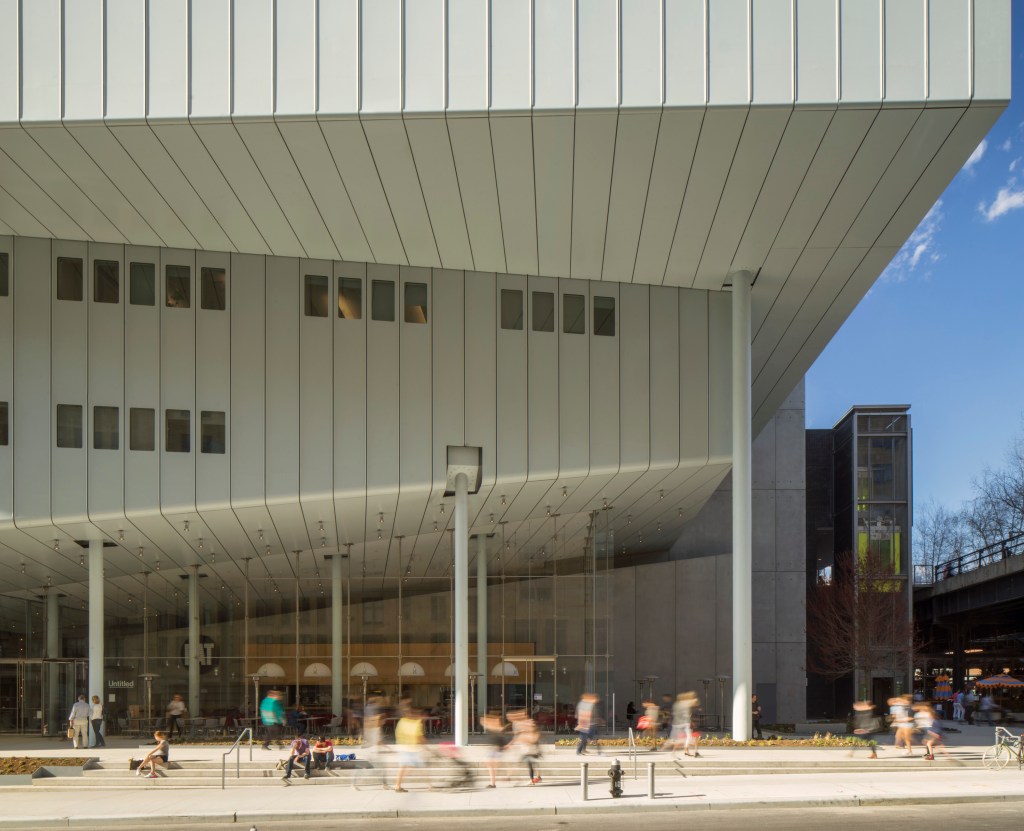 The exterior of a museum building abutting the sidewalk, where crowds are seen walking by, and from which the lobby of the museum can be seen behind glass windows.