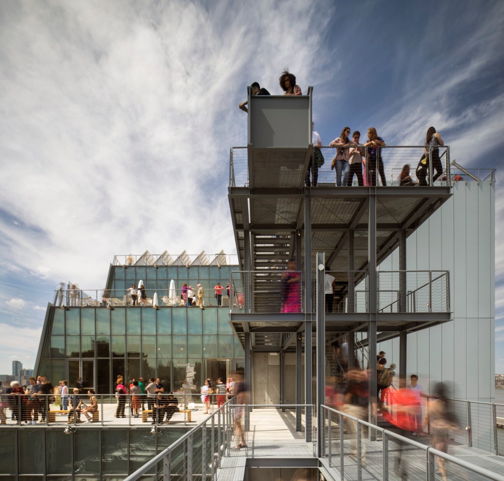 Exterior of a building with glass windows and several levels of balcony terraces where people stand.