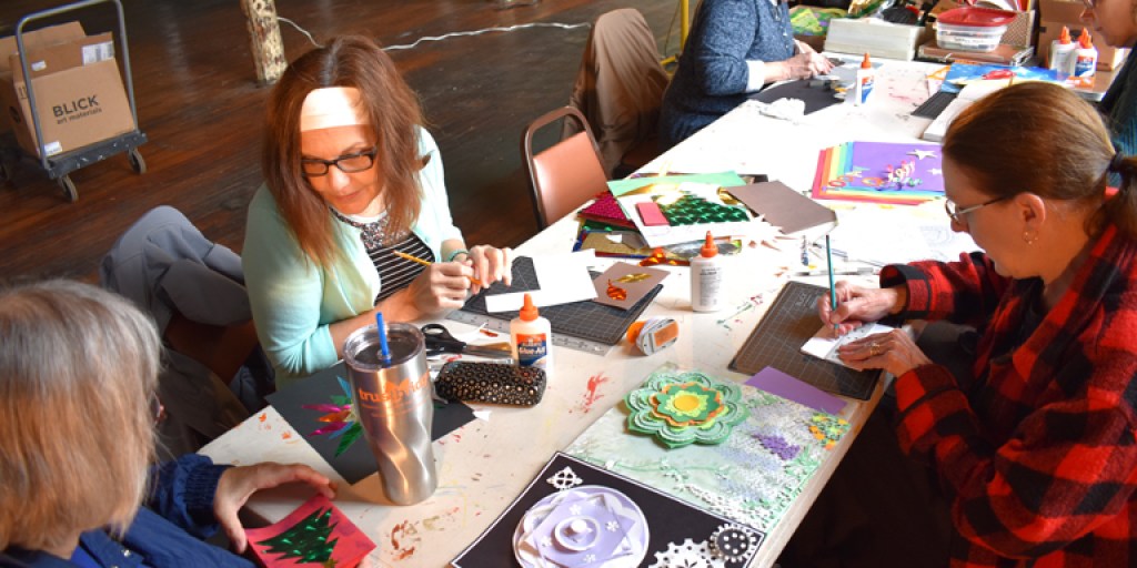 Participants sit around a table working on cut paper projects, with some motifs reminiscent of wheels.