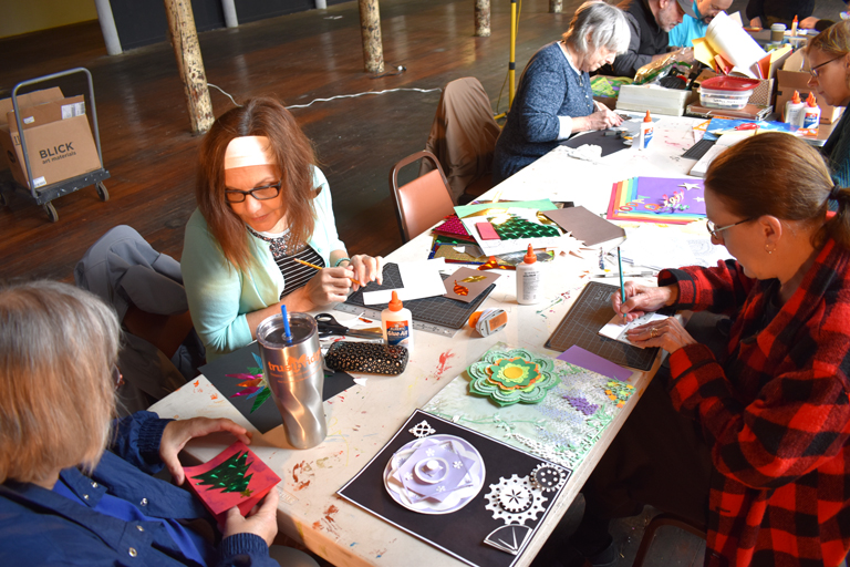 Participants sit around a table working on cut paper projects, with some motifs reminiscent of wheels.