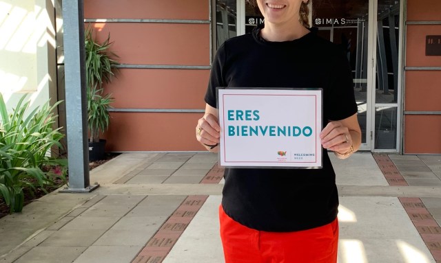 A person stands at the front entrance of the museum holding up a sign that says "ERES BIENVENIDO."