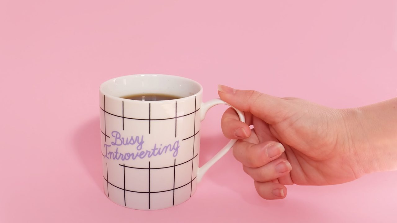 A hand holding a mug that says "busy introverting" against a pink backdrop.