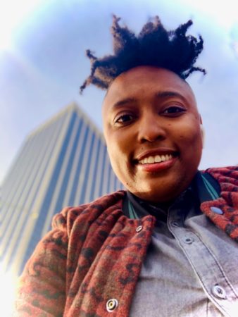 Lanae posing for a selfie with the camera below her and looking up at a tall building and the sky in the background.