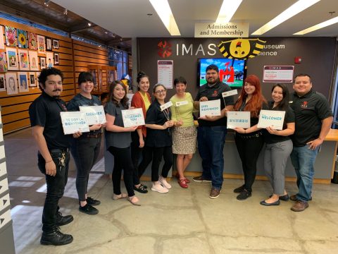 Staff members of the IMAS gather around the admissions and membership desk of the museum, holding up signs that say messages of welcoming in multiple languages.