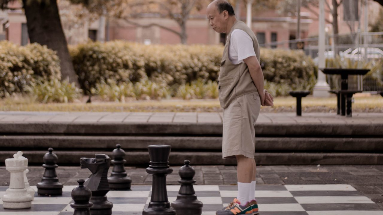 An older person standing on a life-size chessboard in a park.