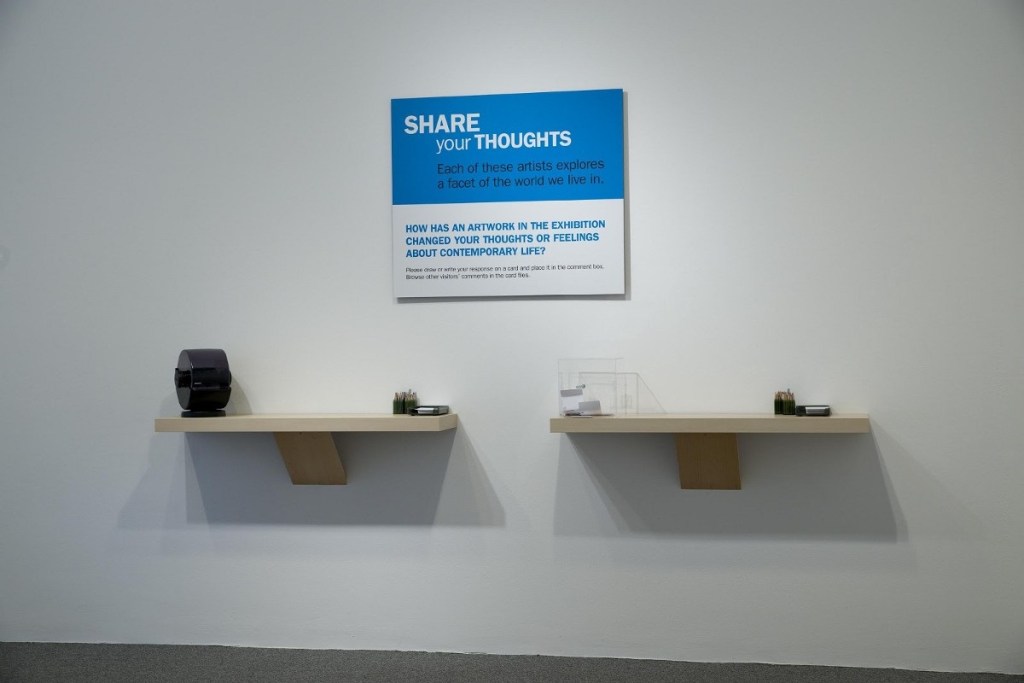 Two floating shelves with note cards, pencils, and a rolodex on them, beneath as sign that says "Share your thoughts: Each of these artists explores a facet of the world we live in. How has an artwork in the exhibition changed your thoughts or feelings about contemporary life?"