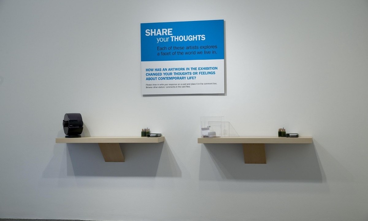 Two floating shelves with note cards, pencils, and a rolodex on them, beneath as sign that says "Share your thoughts: Each of these artists explores a facet of the world we live in. How has an artwork in the exhibition changed your thoughts or feelings about contemporary life?"