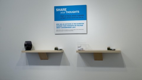 Two floating shelves with note cards, pencils, and a rolodex on them, beneath as sign that says "Share your thoughts: Each of these artists explores a facet of the world we live in. How has an artwork in the exhibition changed your thoughts or feelings about contemporary life?"