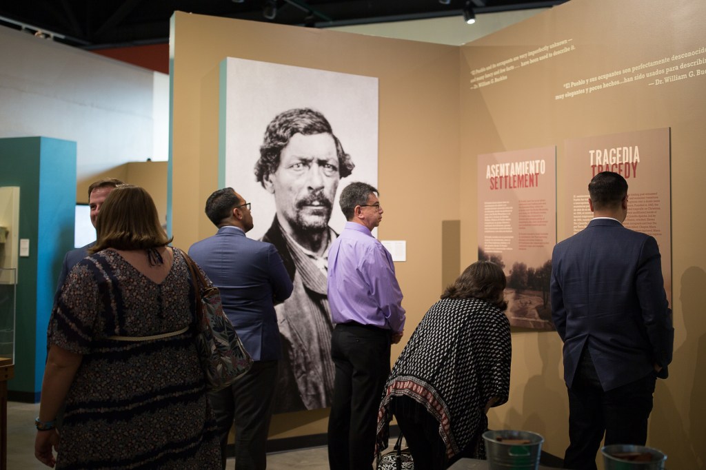 Visitors perusing a gallery with placards reading "settlement" and "tragedy" in English and Spanish.