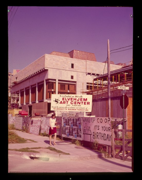 A historical photo of an under-construction building with a plywood wall surrounding it covered in handwritten signs that say things like, "Wake Up Co-op It's Your Birthday!"