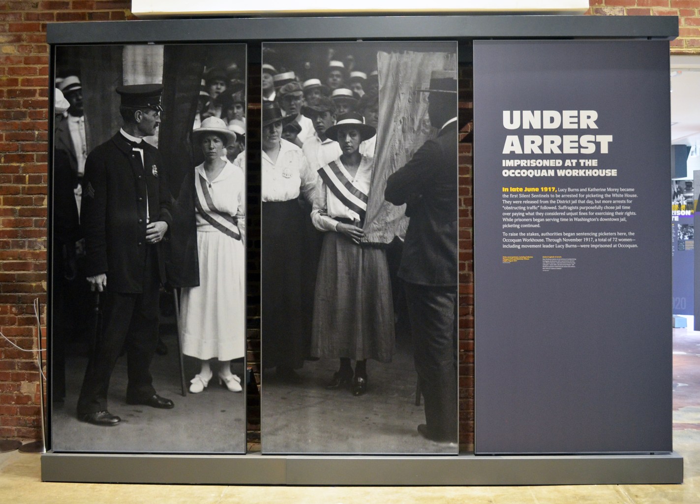 A panel display in a museum showing photos of suffragists facing uniformed men. The text reads "Under Arrest: Imprisoned at the Occoquan Workhouse."