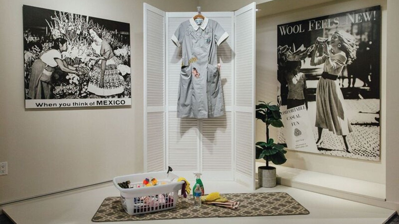 A gray maids uniform hangs from a louvered folding screen with various cleaning products laid out in a laundry basket and on the floor in the Nanny exhibition.