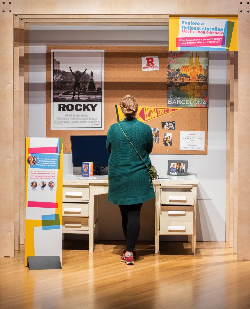 A visitor looking at a display set up to resemble a desk space, with a computer and family photo on the desktop and mementos like posters and college flags on a cork board above. A sign reads, "Explore a fictional storyline about a teen overdose."
