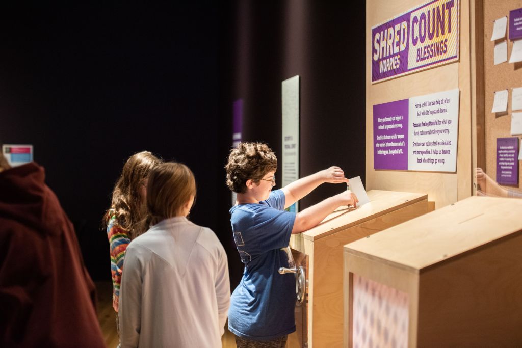 Children standing in front a wooden contraption with a slot in it, under a sign that says "Shred Worries, Count Blessings." One child places a sheet of paper into the slot.