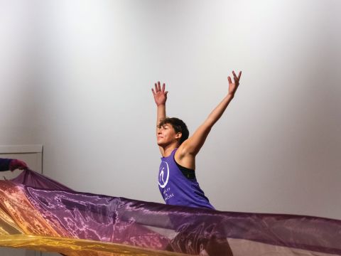 A person stands behind a colorful sheer piece of fabric that is blowing in the wind with their arms outstretched as though taking flight.