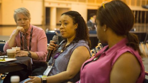 Two people listening to another person speaking into a microphone.