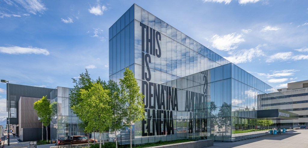 A glass building facade with large letters reading "This is Dena'ina Elnena"